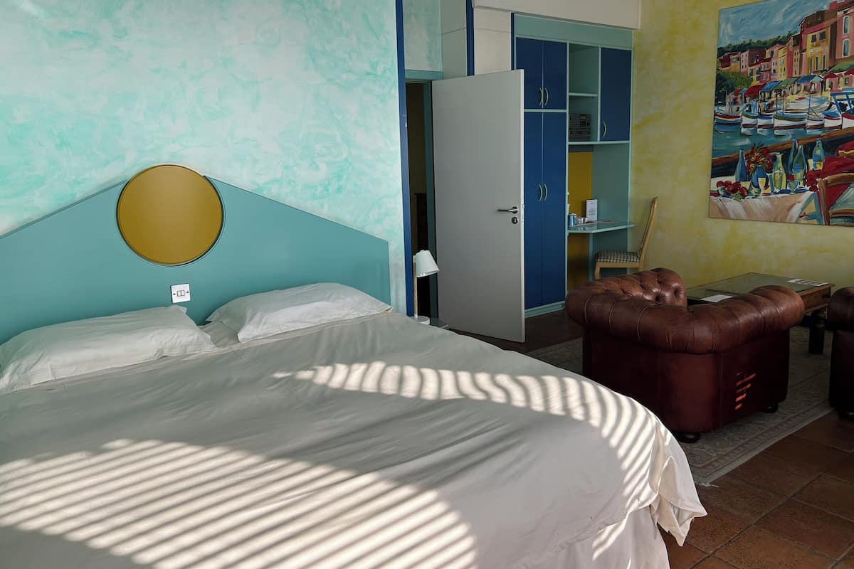 A hotel room with a bed, a round mirror, a wardrobe, a seating area with a brown leather sofa, and a painting on the wall.
