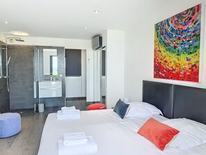 A modern hotel room with a large bed, a bathroom with a shower, a sink, and a mirror. There is a colorful abstract painting on the wall.