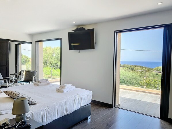 A modern hotel room with a large bed, a flat-screen TV, and a balcony with a view of the sea.