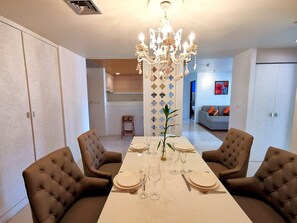 A dining room with a long table set for four, surrounded by tufted chairs, a chandelier, and a decorative wall panel.