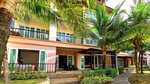 A two-story building with a covered outdoor seating area, palm trees, and a paved walkway. - Apartment with private jacuzzi and pool access. Just 800 meters to the beach. (Phuket)