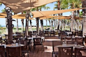 Een terras met tafels en stoelen, beschaduwd door grote parasols, omgeven door palmbomen en uitzicht op het strand.