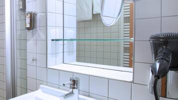 Ein Badezimmer mit weißem Waschbecken, einer Duschkabine aus Glas, einem Spiegel und einem an der Wand befestigten Föhn.