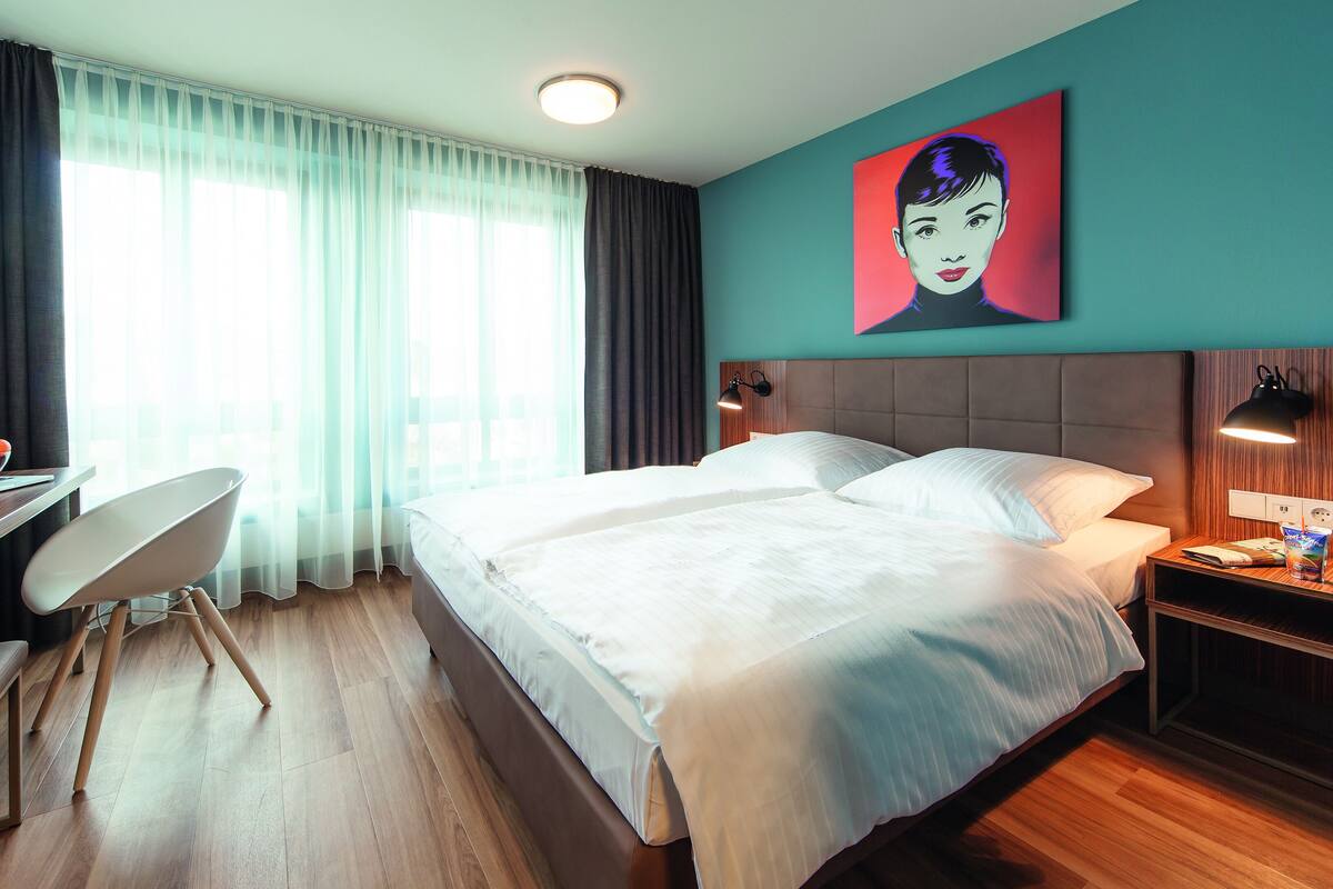 A hotel room with a bed, desk, chair, and a painting on the wall.