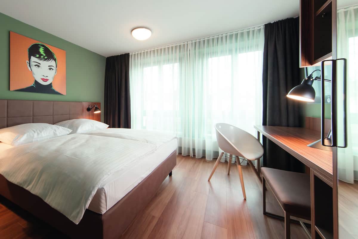 A hotel room with a large bed, a desk with a chair, a wall-mounted lamp, and a window with curtains.