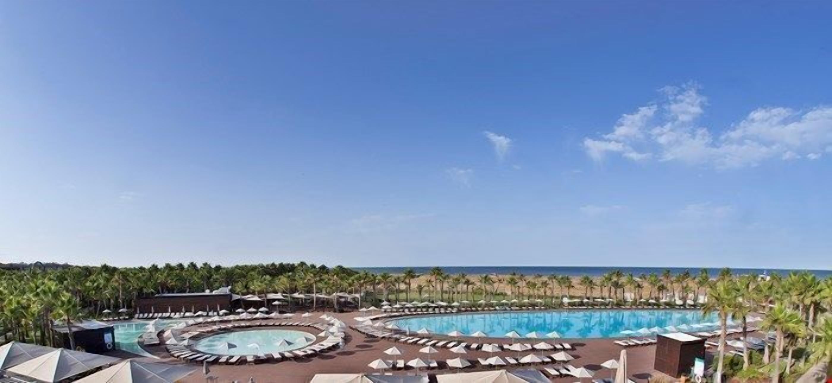 Een resort met meerdere zwembaden, ligbedden en parasols, omringd door palmbomen en uitzicht op de oceaan.