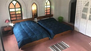 Una habitación con dos camas, cabecero de madera, mesita de noche con lámpara y tres ventanas arqueadas con cortinas.