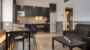Una cocina moderna con armarios oscuros, un microondas y un refrigerador. Hay un comedor con mesa y sillas, y un sofá en la sala.