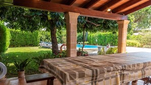 Une terrasse extérieure couverte, avec une table dressée pour le repas, entourée d’une végétation luxuriante et une piscine en arrière-plan.