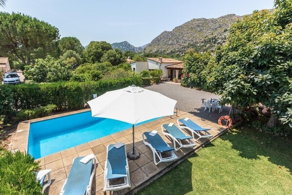 A swimming pool with lounge chairs and a large umbrella, surrounded by greenery and a mountainous backdrop.
