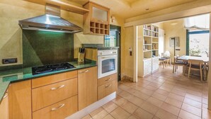 A kitchen with a stove, oven, and wooden cabinets. There is a dining area with a table and chairs visible through an open doorway. - Oliver Conti (House, Capmany) (Capmany)