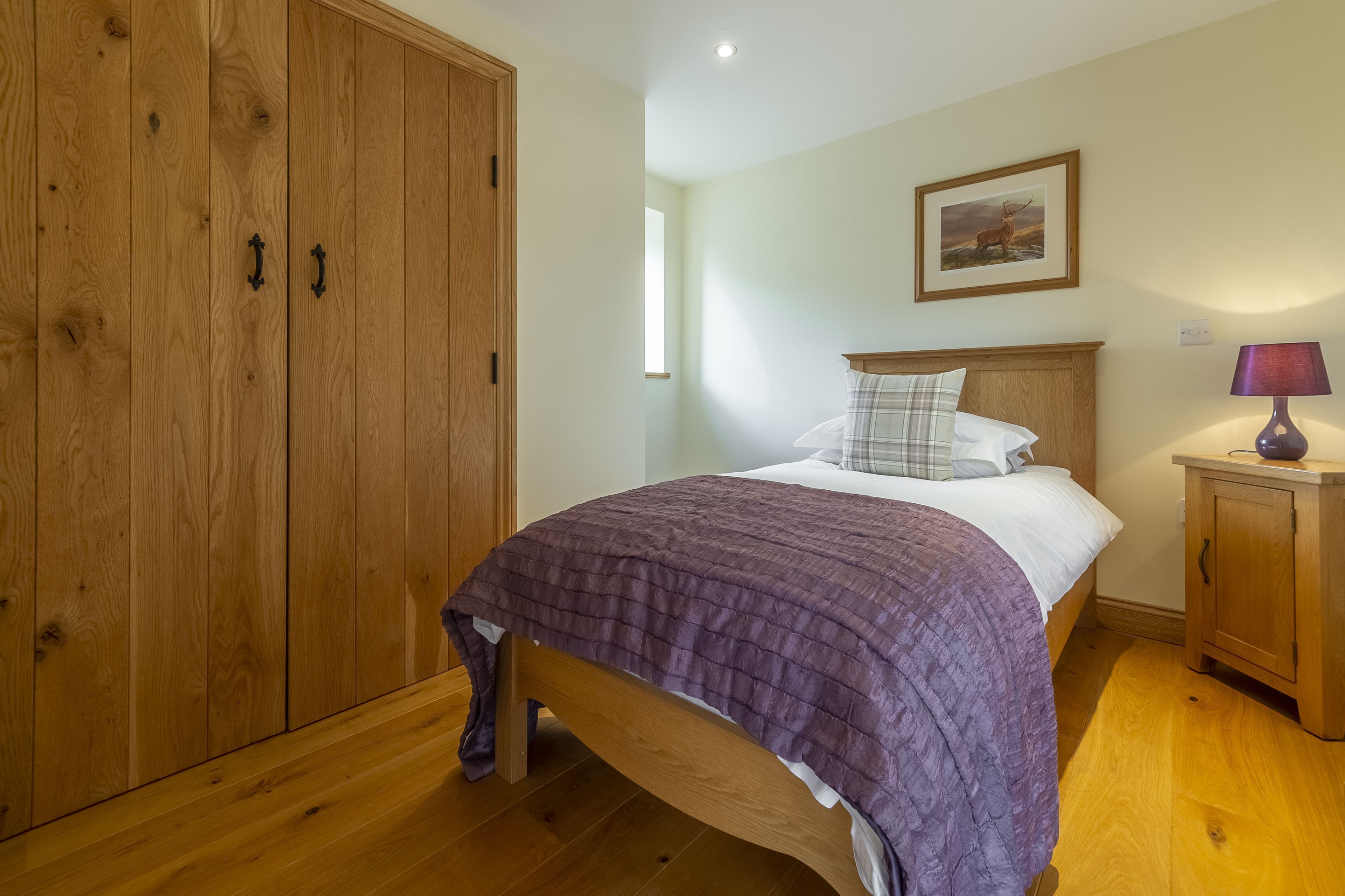 A bedroom with a wooden wardrobe, a bed with a purple blanket, a nightstand with a lamp, and a framed picture on the wall.