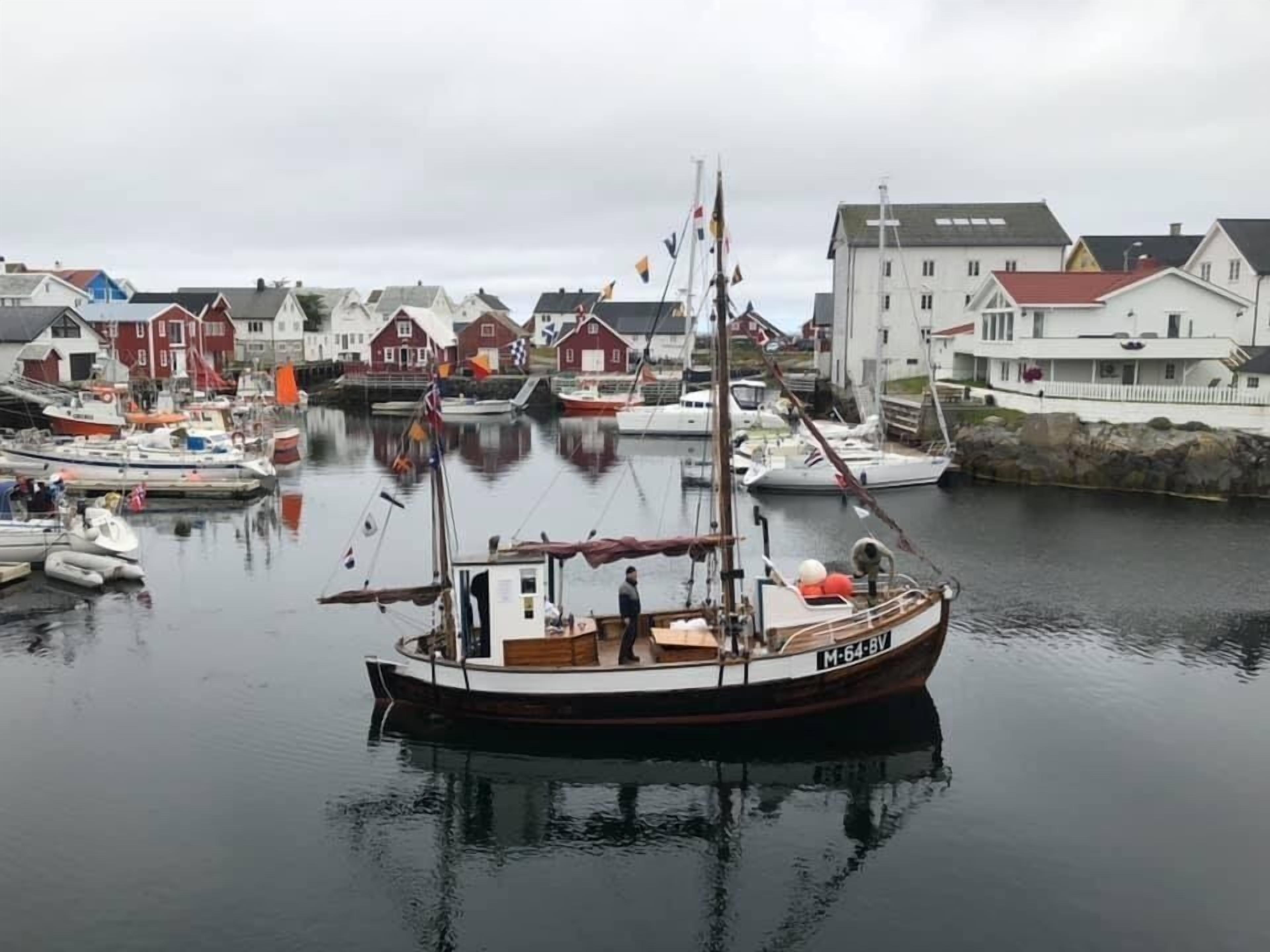 A harbor with boats docked, a small cabin boat with a cabin and a flag, and buildings in the background.