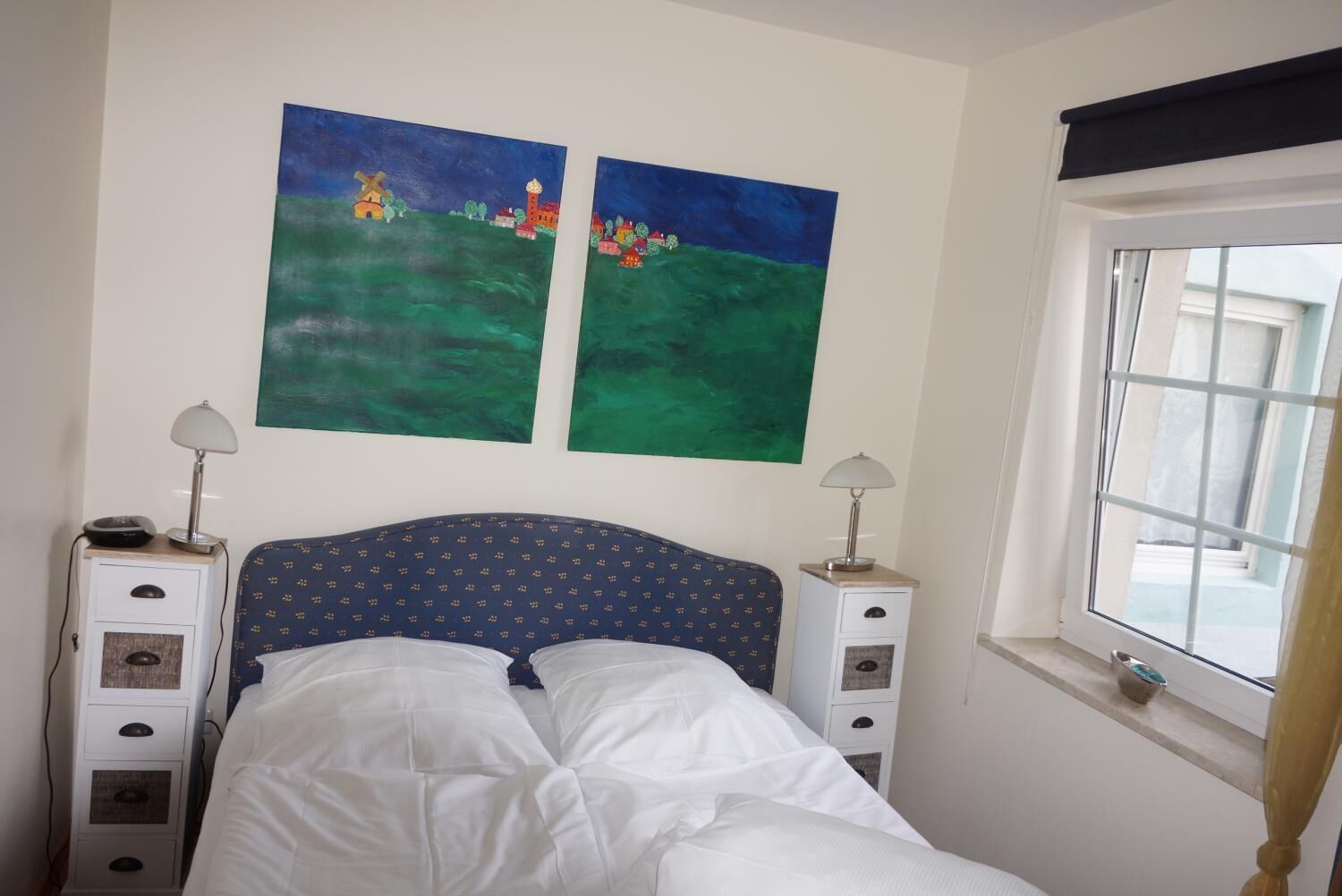 Quarto com cama, duas mesas de cabeceira, uma janela e duas pinturas na parede.