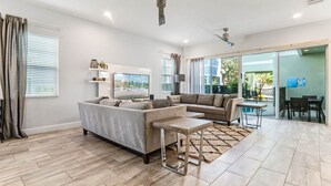 A modern living room with a sectional sofa, a coffee table, and a TV. There is a sliding glass door leading to a patio area. - EV_1472 - 5 Bed 5.5 Bath Villa with Screened-In Private Pool, Kids Room (Kissimmee)
