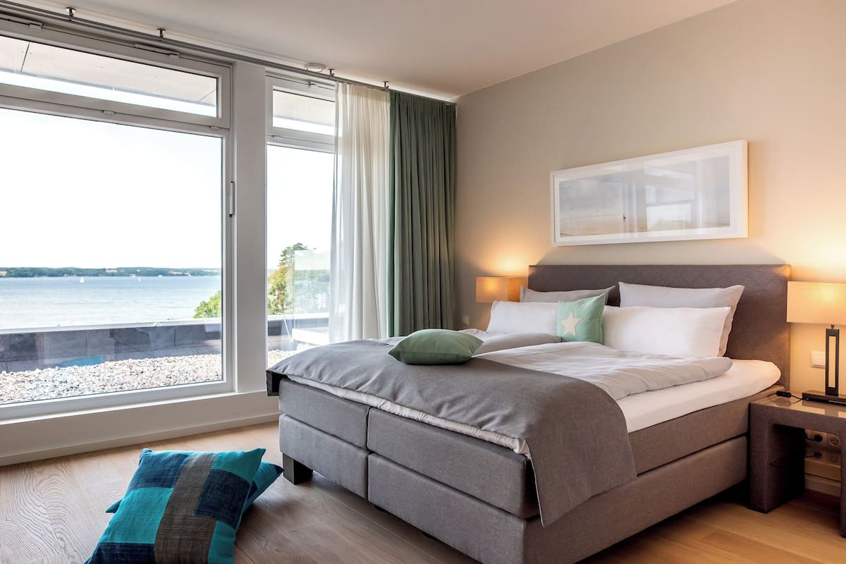 Ein Schlafzimmer mit einem Bett, einem Nachttisch, einem Fenster mit Vorhängen und Blick auf das Wasser.