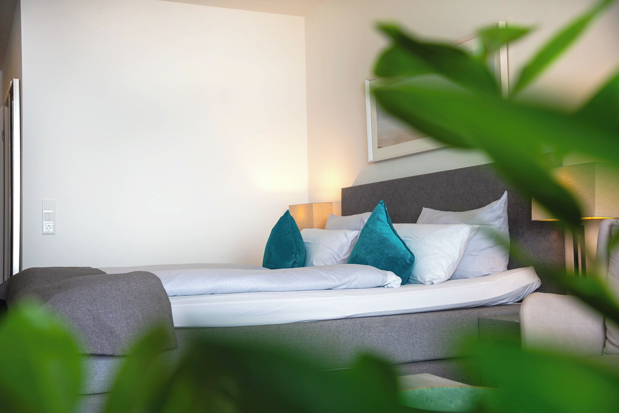 Una cama bien hecha con almohadas blancas y azul turquesa, un cabecero gris y una lámpara de noche.