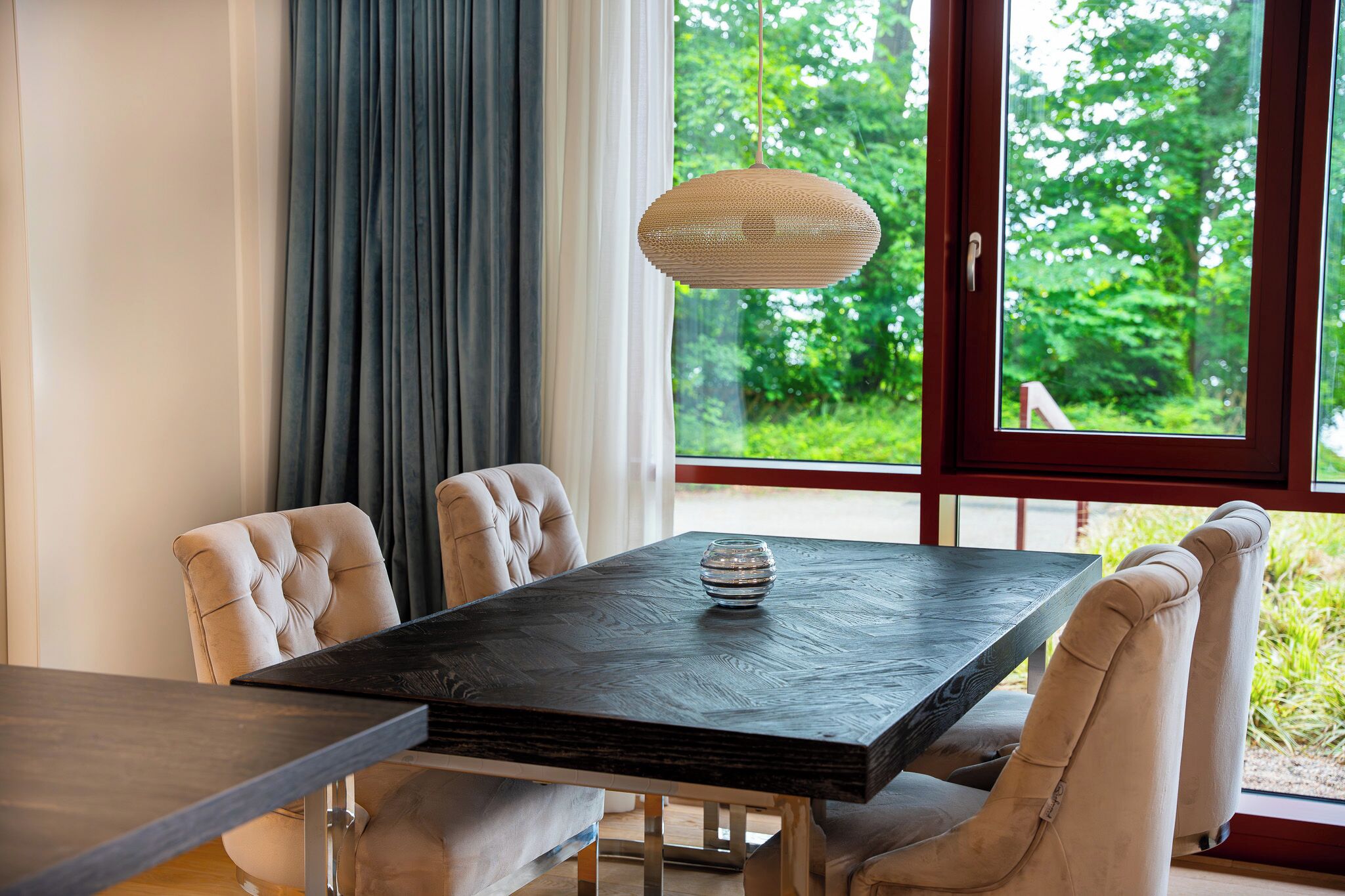 A dining table with chairs, a hanging light fixture, and a view of greenery through a window.
