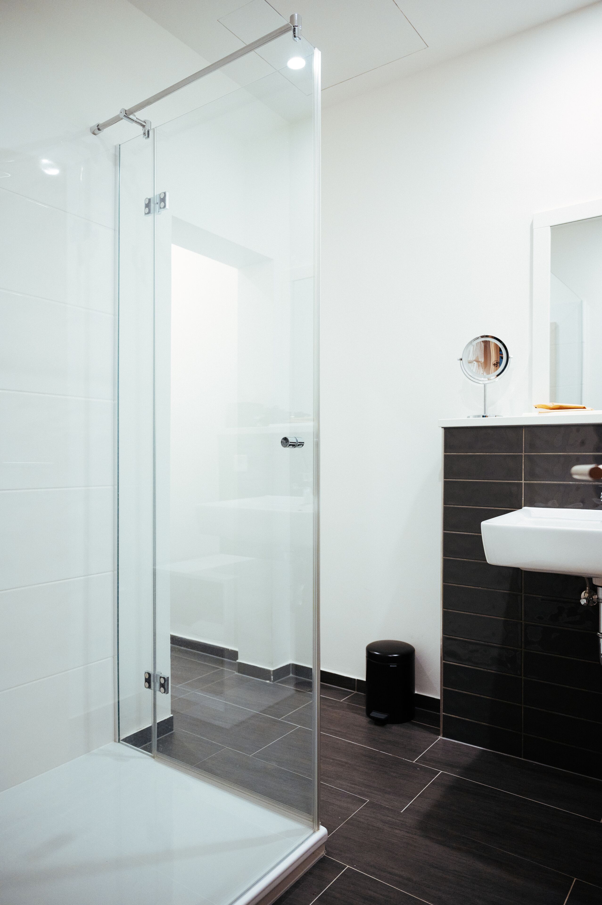 Un baño moderno con cabina de ducha de vidrio, un lavamanos blanco y una pared revestida de azulejos negros.