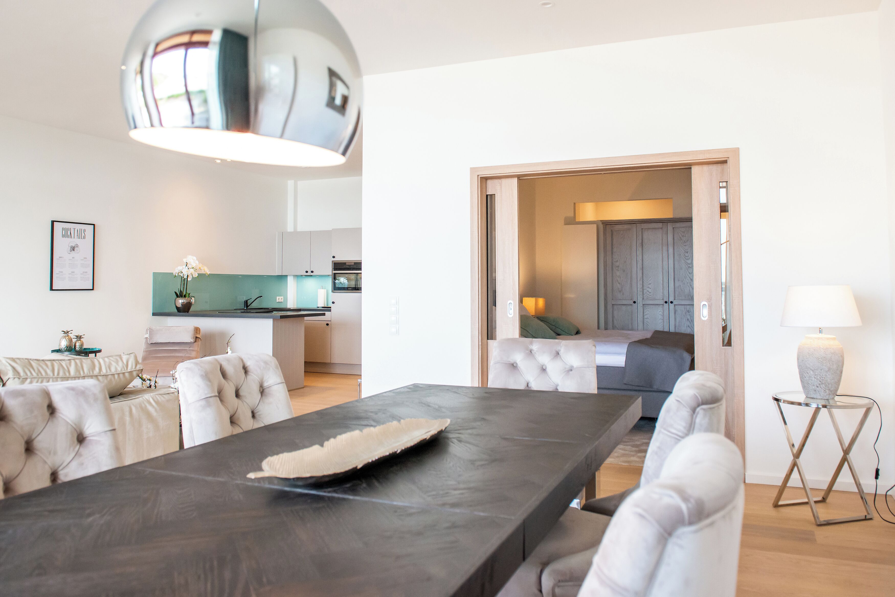 A modern living room with a dark wooden dining table, white upholstered chairs, a tufted sofa, a large mirror, and a kitchen in the background.