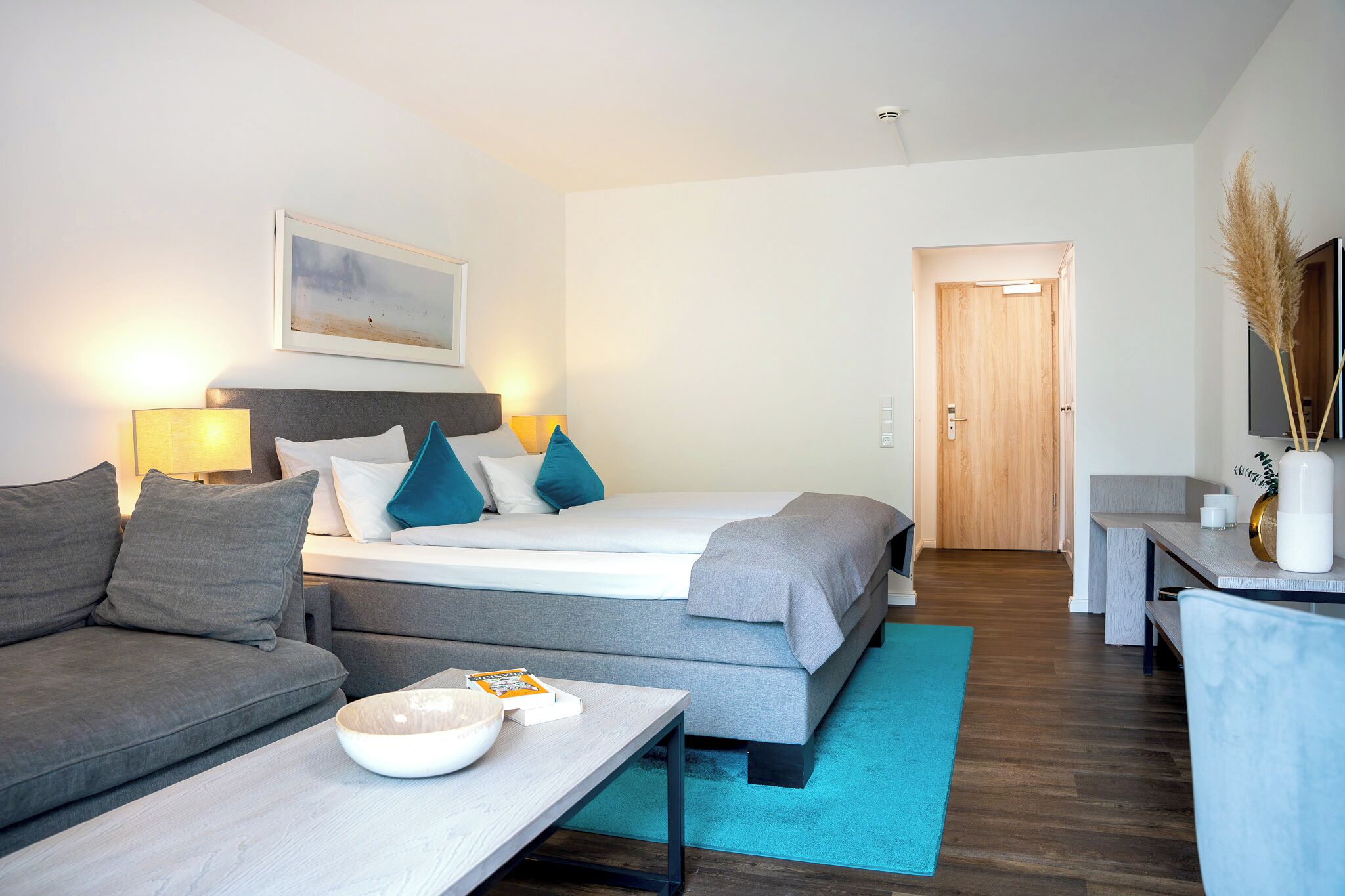 Una habitación de hotel moderna con cama, sofá, mesita de noche, lámpara y un cuadro en la pared.