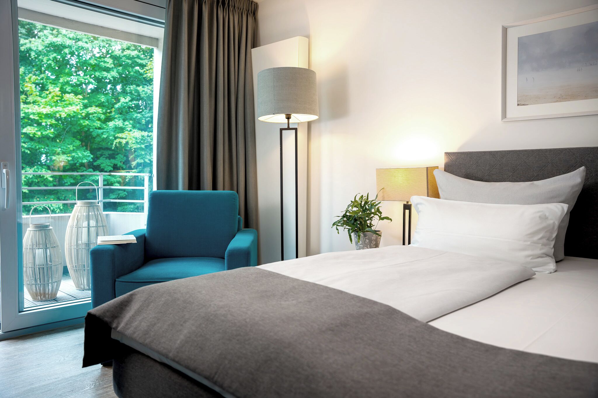 Ein modernes Hotelzimmer mit einem großen Bett, einem blauen Sessel, einer Stehlampe und Blick ins Grüne.