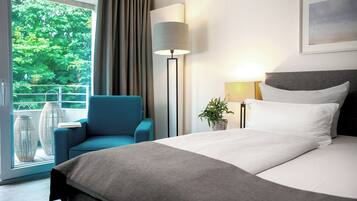 Ein modernes Hotelzimmer mit einem großen Bett, einem blauen Sessel, einer Stehlampe und Blick ins Grüne.
