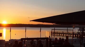 A sunset view from a waterfront restaurant with tables and chairs, a pier in the background, and a calm body of water.