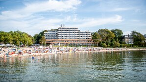 Ein Hotel am Wasserufer mit mehreren Etagen, ein Strandbereich inklusive Sonnenschirmen und Liegen, und ein ruhiger Fluss im Vordergrund.