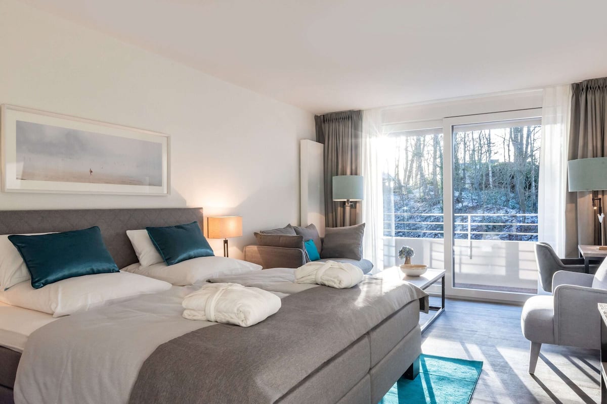 Ein Hotelzimmer mit einem großen Bett, einem Schreibtisch, einem Sessel und Blick ins Freie, sowie einem Bild an der Wand.