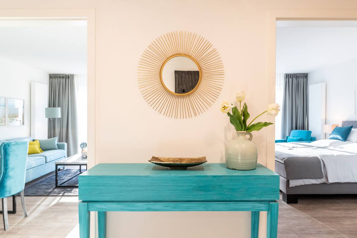 Ein modernes Hotelzimmer mit einem türkisfarbenen Konsolentisch, einem runden Spiegel mit strahlender Umrandung und einer Vase mit weißen Blumen.