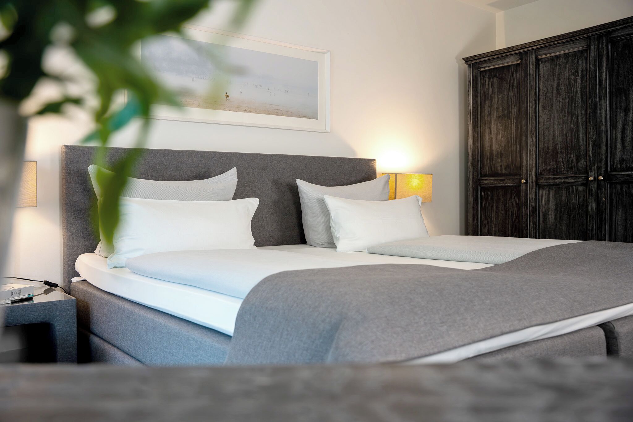Una cama bien hecha con ropa de cama blanca y gris, una mesita de noche con lámpara y un armario de madera oscura.