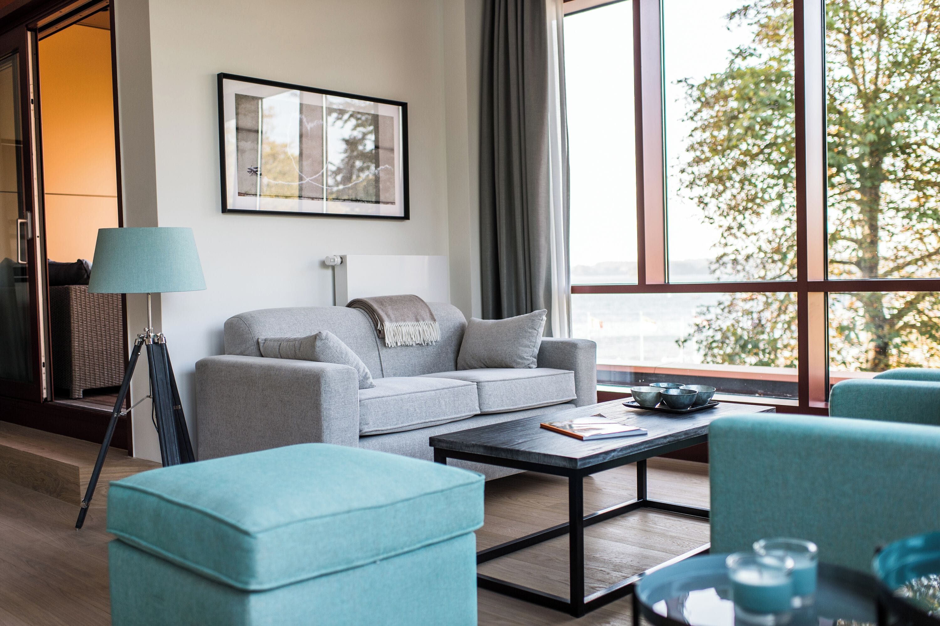 A modern living room with a sofa, ottoman, and coffee table, a framed picture on the wall, and a large window with a view.
