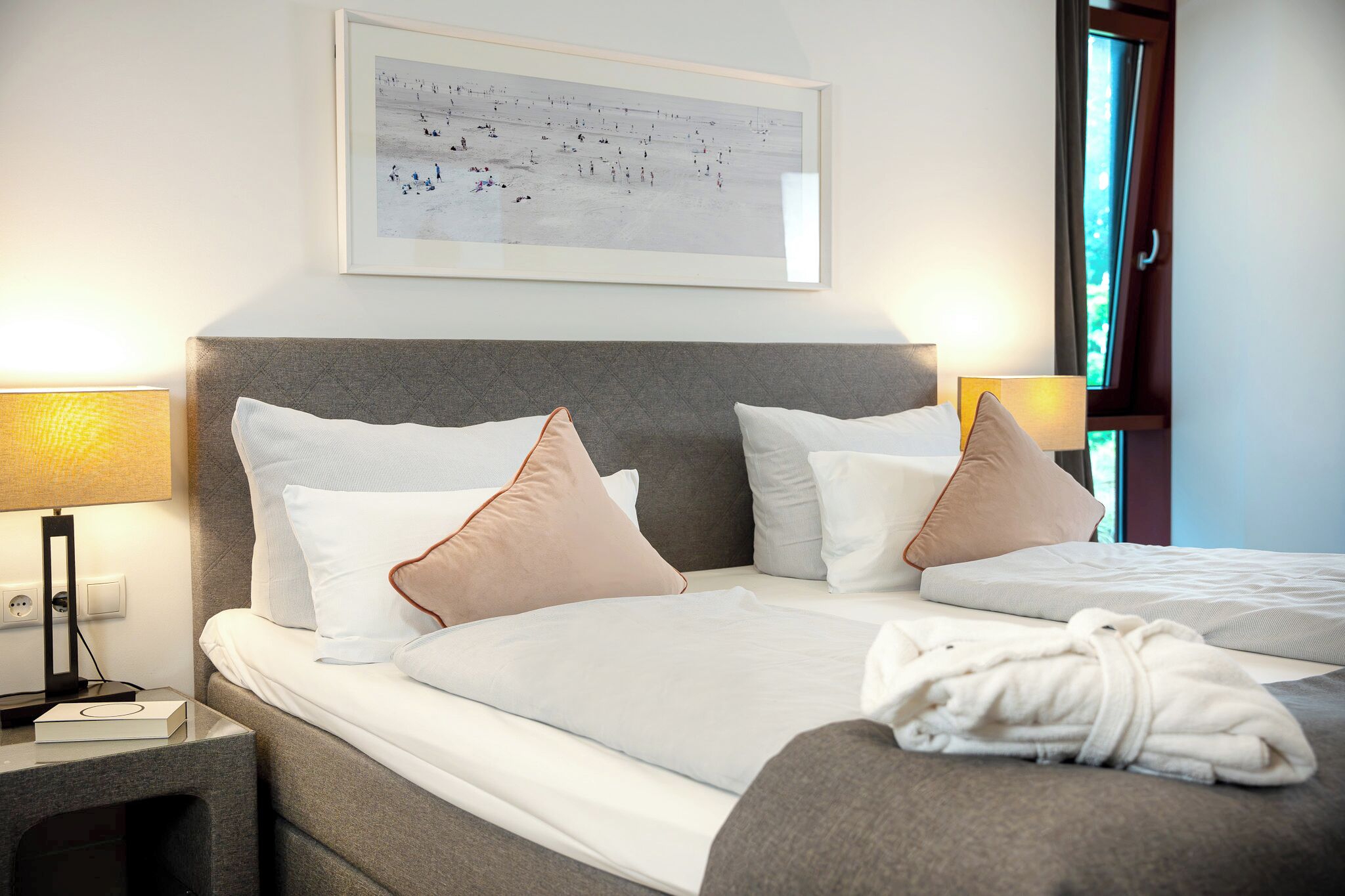 A hotel room with a bed, pillows, a bedside table with a lamp, a framed picture on the wall, and a window.