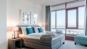 Ein modernes Hotelzimmer mit einem großen Bett, einem Nachttisch, einem Fenster mit Meerblick und Kunstwerken an der Wand.