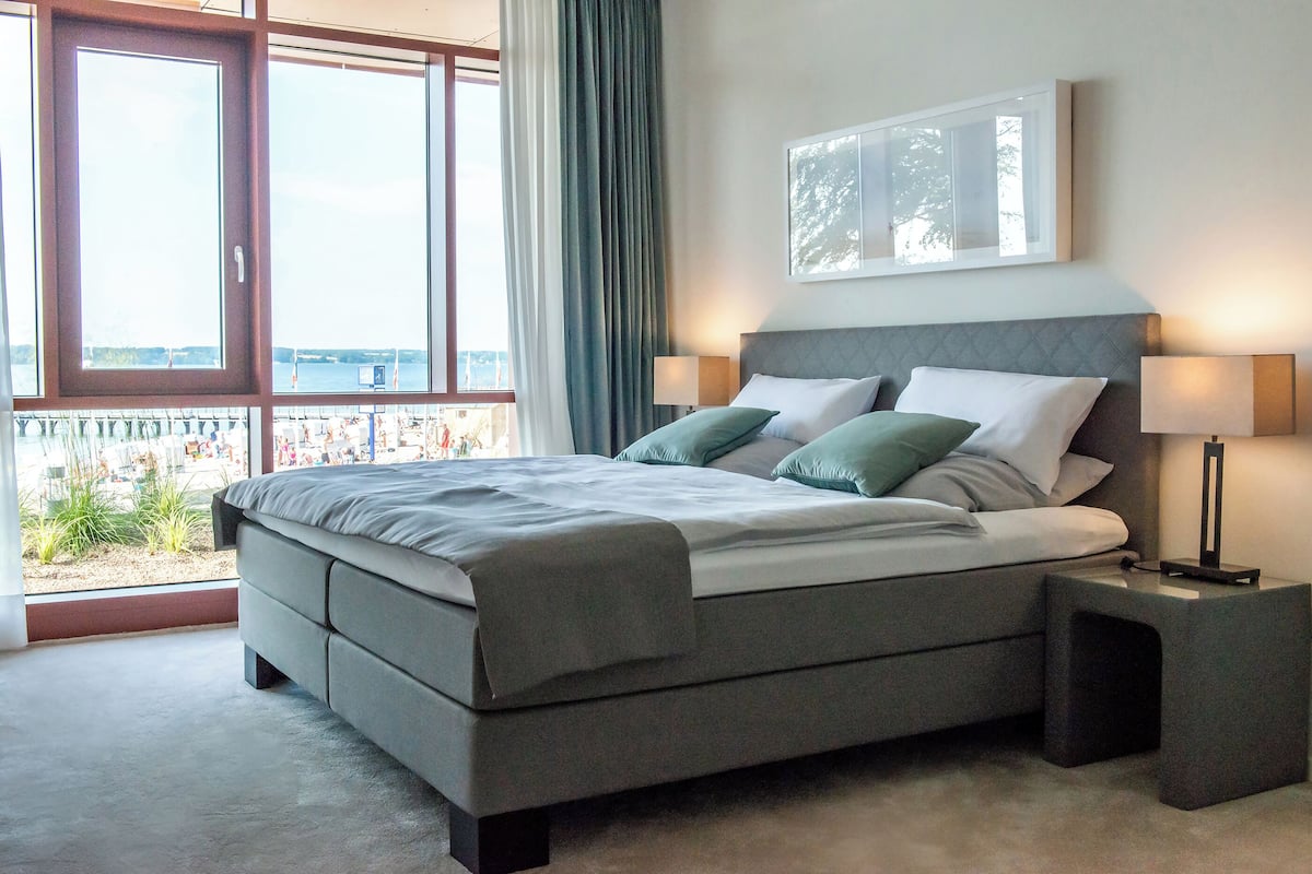 Ein modernes Schlafzimmer mit einem großen Bett, zwei Nachttischen, einem Fenster mit Blick und einem gerahmten Bild an der Wand.