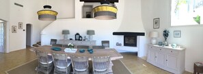 Uma área de refeições com uma grande mesa de madeira, cadeiras de vime e duas luminárias pendentes com cúpulas estampadas.