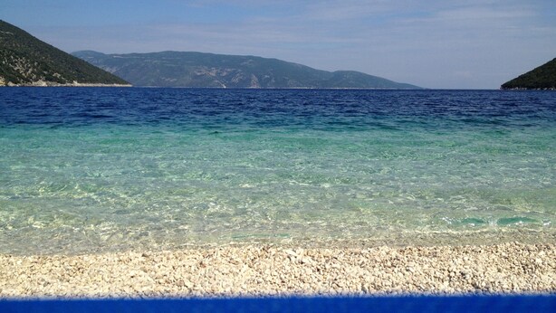 On aperçoit clairement une plage de galets qui s’étend jusqu’à une mer calme, avec des montagnes qui se dessinent à l’horizon.