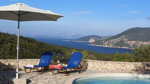 Un espace piscine agrémenté d’une terrasse en pierre, de deux chaises longues bleues, d’un parasol blanc et offrant une vue sur la mer et les montagnes.