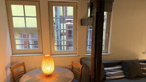 Una habitación acogedora con una mesa redonda de madera, dos sillas, una lámpara de pie, un sofá y dos ventanas que dan a vistas de edificios.