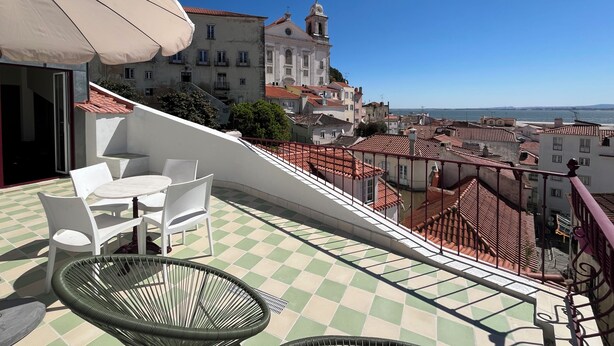 Una terraza con suelo a cuadros, sillas blancas y una silla verde, con vistas a un paisaje urbano con edificios y cielo despejado.