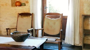 Een rustieke kamer met twee houten stoelen, een tafel met een kom, en een klein schap met fruit en een bak.