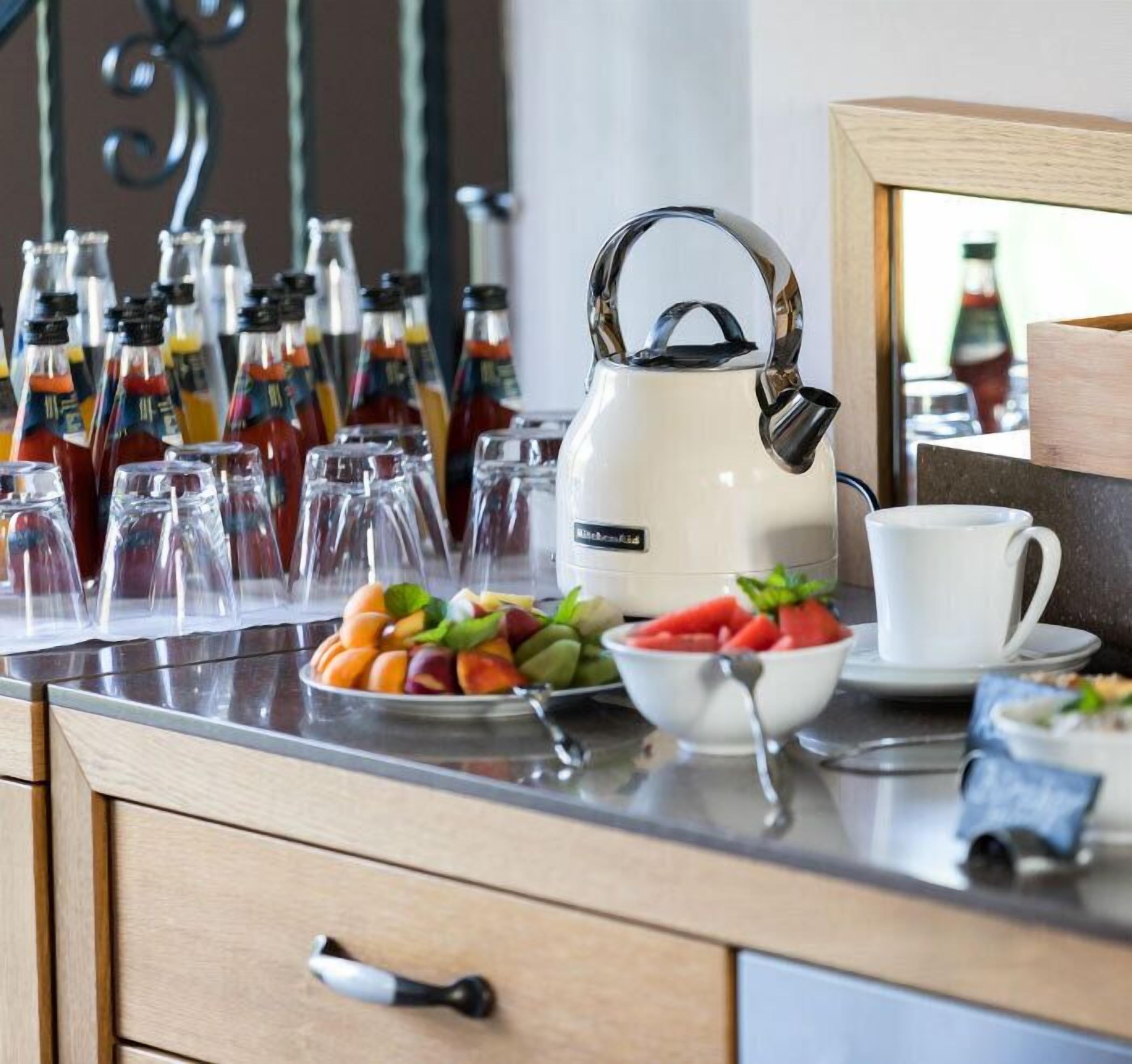 A kitchen counter with a white kettle, a bowl of fruit, a cup, and several bottles of beverages.