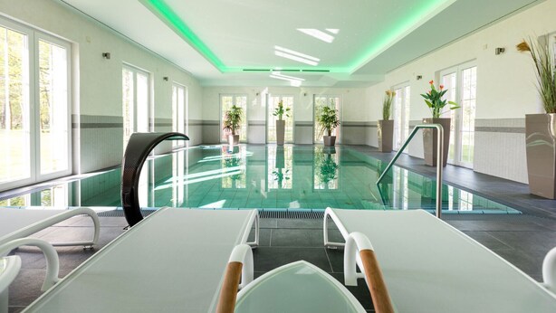A modern indoor swimming pool with clear glass walls, a central pool area, and multiple lounge chairs.