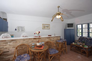 A kitchen with a stone backsplash, wooden chairs, a dining table, a ceiling fan, and a TV on the wall. - Manos Syros (Syros)