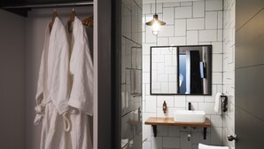 A modern bathroom with a white tiled wall, a wooden vanity, a large mirror, and two white bathrobes hanging on a rack. - The Pop Hotels (Santiago)