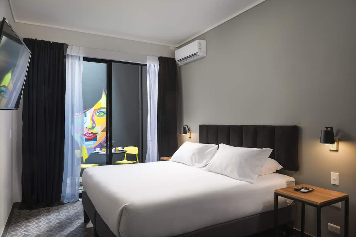A hotel room with a bed, a TV, a wall-mounted air conditioner, a nightstand, and a chair.