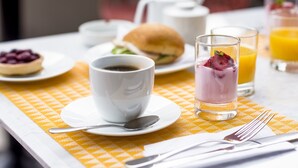 A breakfast setting with a cup of coffee, a glass of orange juice, a sandwich, and a dessert on a checkered tablecloth. - The Pop Hotels (Santiago)