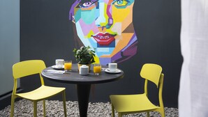 A dining area with a round table, two yellow chairs, a potted plant, and a colorful mural on the wall. - The Pop Hotels (Santiago)