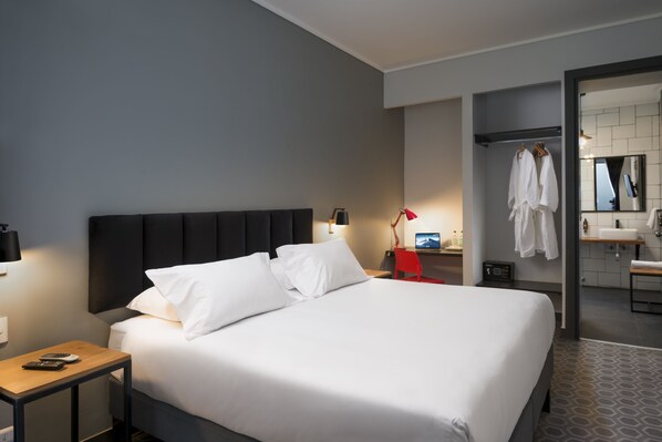 A modern hotel room with a large bed, bedside tables, a desk, and a bathroom visible through an open door.